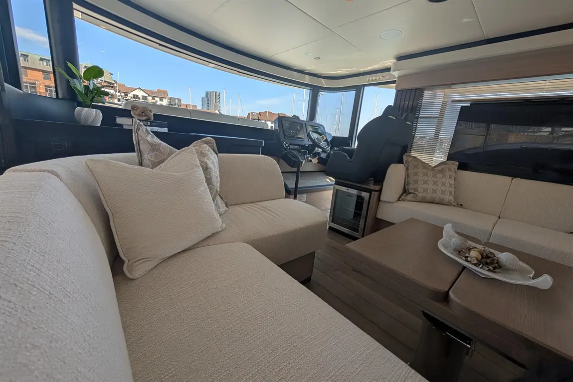 Stylish boat interior featuring a beige sofa, decorative pillows, a coffee table with shells, and a modern helm with views of the marina.